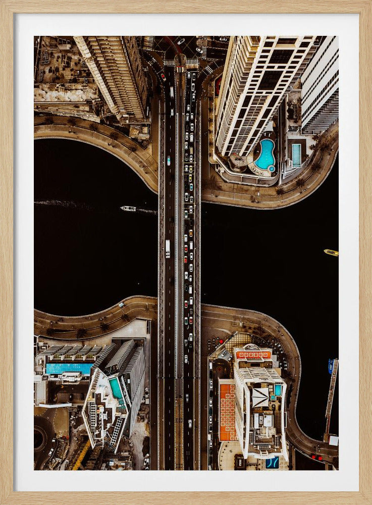 A symmetrical, top-down aerial photograph of a busy city bridge crossing dark water, flanked by towering skyscrapers with bright blue rooftop swimming pools. Print