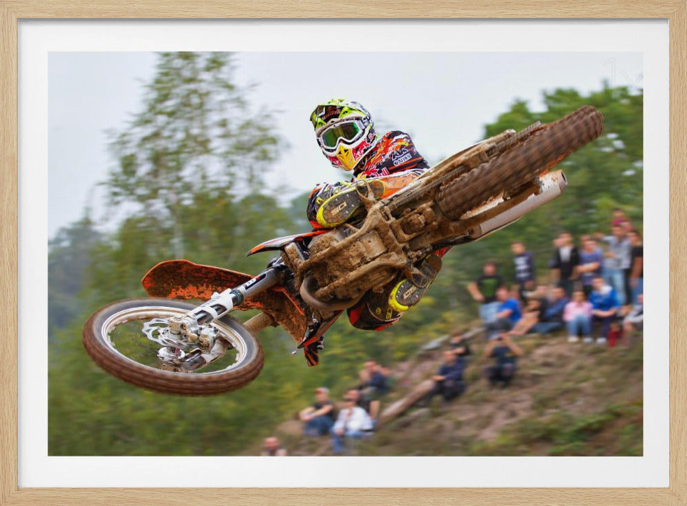 A dynamic action shot of a motocross racer in mid-air, leaning into a turn with their muddy orange dirt bike. The rider wears a colorful helmet and racing gear, with a blurred background of green trees and spectators watching from a dirt hill. The photo is enclosed in a silver frame. Decor