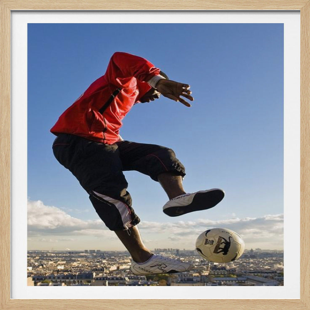 A dynamic low-angle shot of a person in a red jacket and black pants skillfully juggling a soccer ball, suspended in mid-air high above a sprawling city skyline under a clear blue sky, all enclosed within a light wood frame. Artwork
