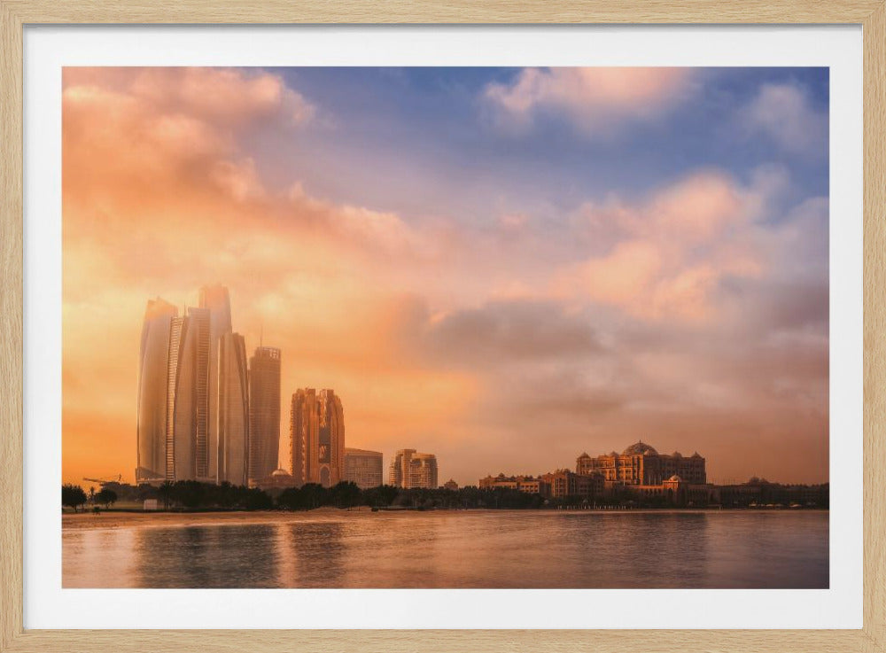 A framed photograph of the Abu Dhabi city skyline at sunset, featuring modern skyscrapers and the Emirates Palace hotel glowing in warm, orange light. The buildings are reflected in the calm water of the foreground, under a sky with soft orange and blue clouds. Poster
