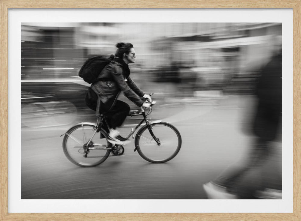 A dynamic black and white photograph capturing a person in profile as they cycle swiftly through a city. The background is a motion blur of urban scenery, highlighting the cyclist's speed. The individual wears a leather jacket, sunglasses, a backpack, and has their hair tied up. The artwork is enclosed in a silver frame. Wall Art