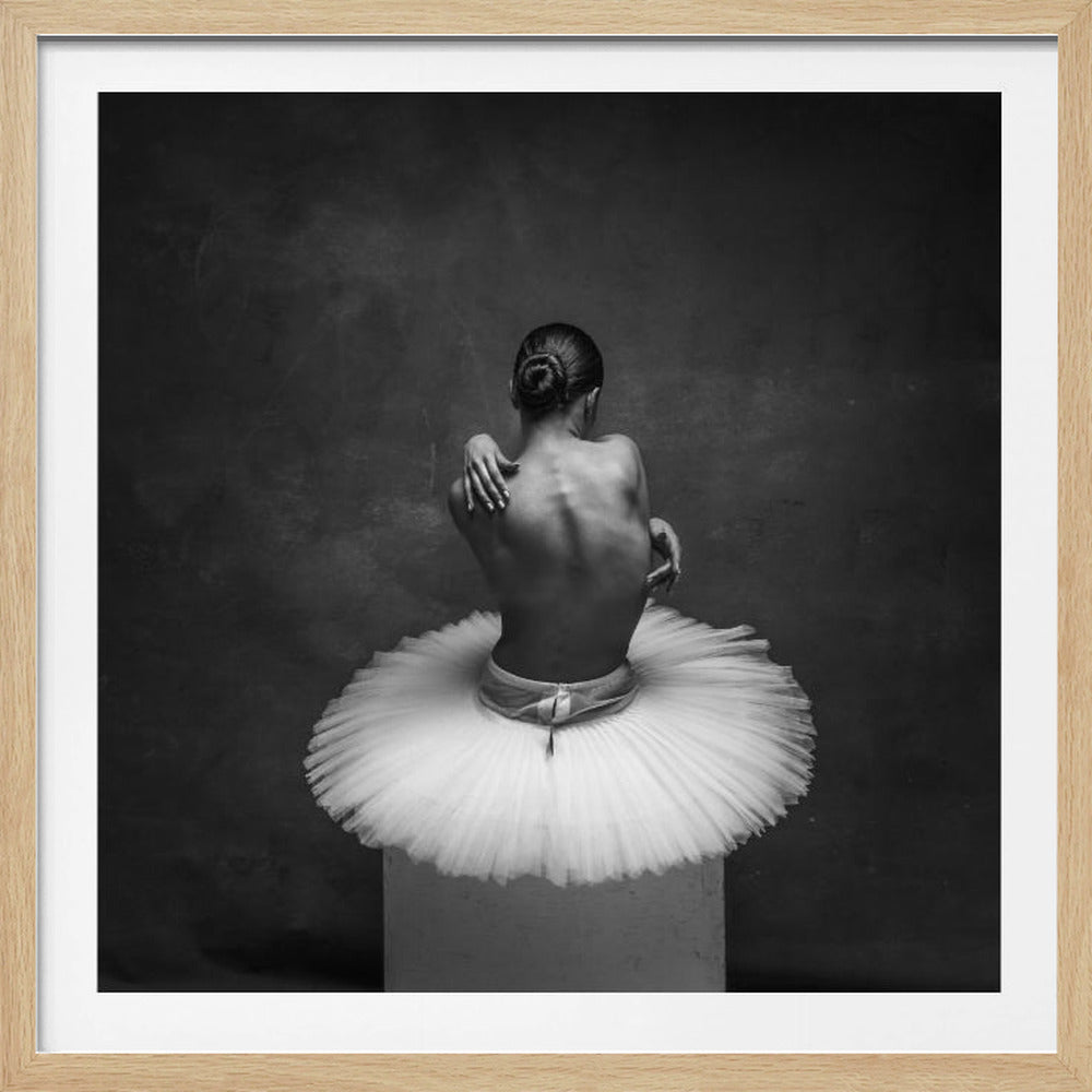 A framed, black and white photograph of a ballerina seen from behind. She is sitting on a white block, wearing a voluminous white tutu, with her hair in a neat bun. Her arms are wrapped around her bare shoulders, highlighting the muscles of her back against a dark, moody, textured background. Decor