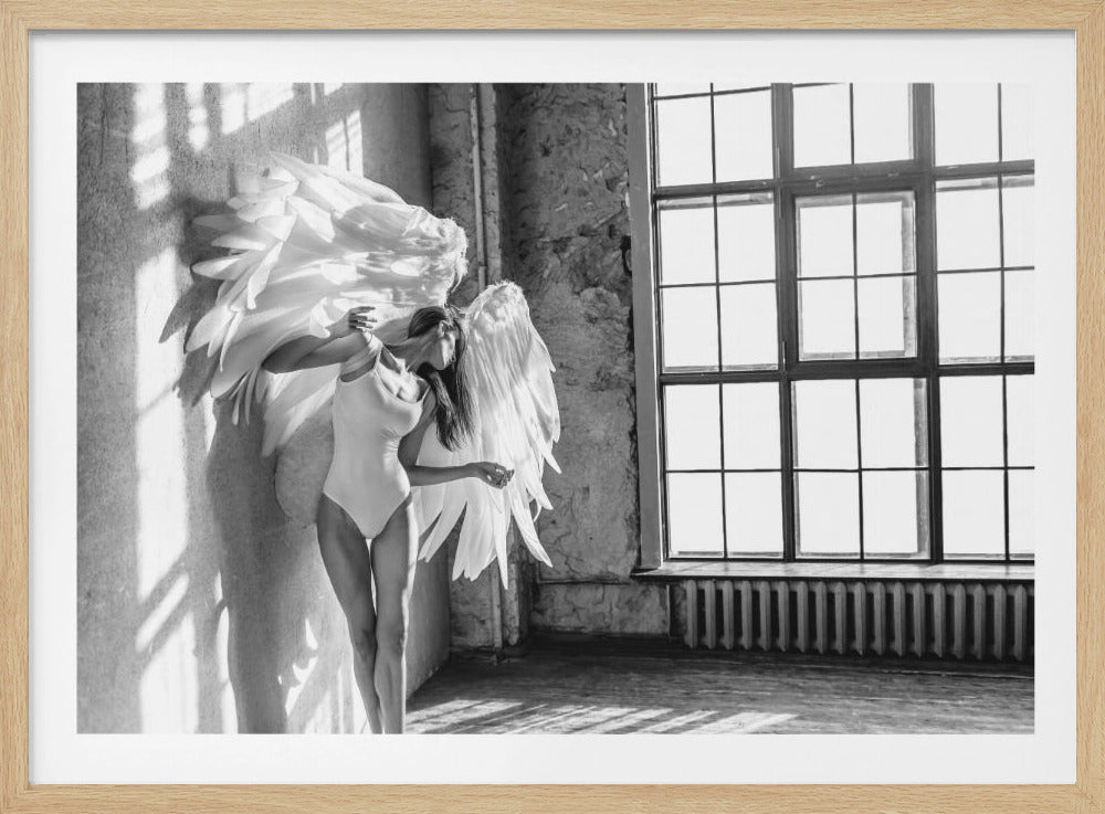 A black and white photograph of a slender woman wearing a white leotard and large, feathery angel wings. She stands gracefully against a rough, sun-drenched wall in an industrial-style room, with a large paned window to the right casting strong light and shadows. Print