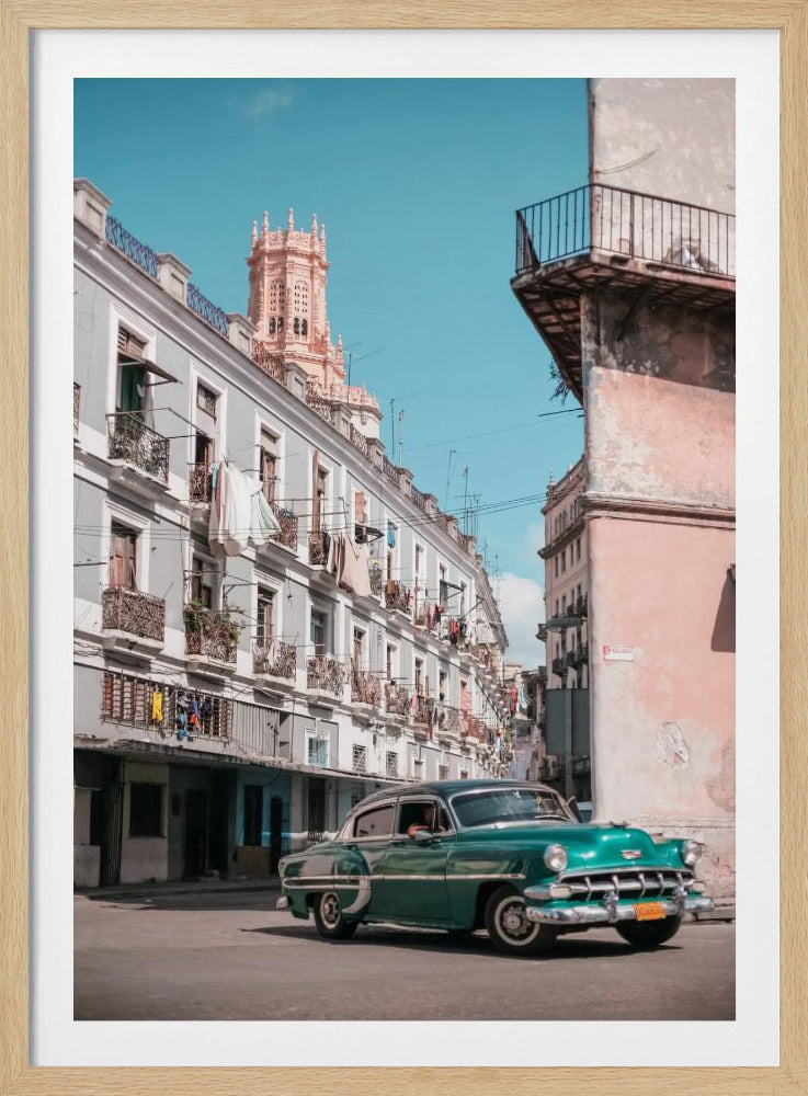 A vintage teal-green classic car sits on a sunny street in a historic city, possibly Havana, Cuba. Weathered apartment buildings with intricate balconies and hanging laundry line the street, with a peach-colored church tower visible in the background against a clear turquoise sky. Poster