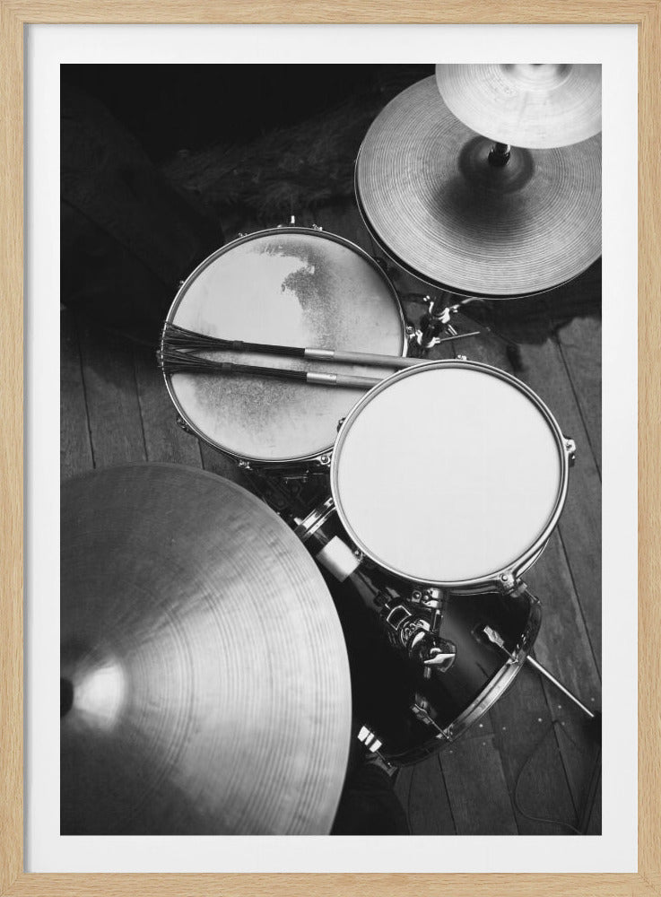 A high-angle, black and white photograph of a drum kit. The focus is on a snare drum with wire brushes resting on it, a tom, and several shiny cymbals, all set on a dark wooden floor. Print