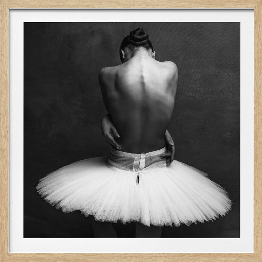 A black and white photograph of a ballerina with her back to the camera. Her hair is in a bun and she's wearing a white tutu. Her hands are clasped behind her at her waist, accentuating the musculature and curve of her spine against a dark, textured background. Artwork