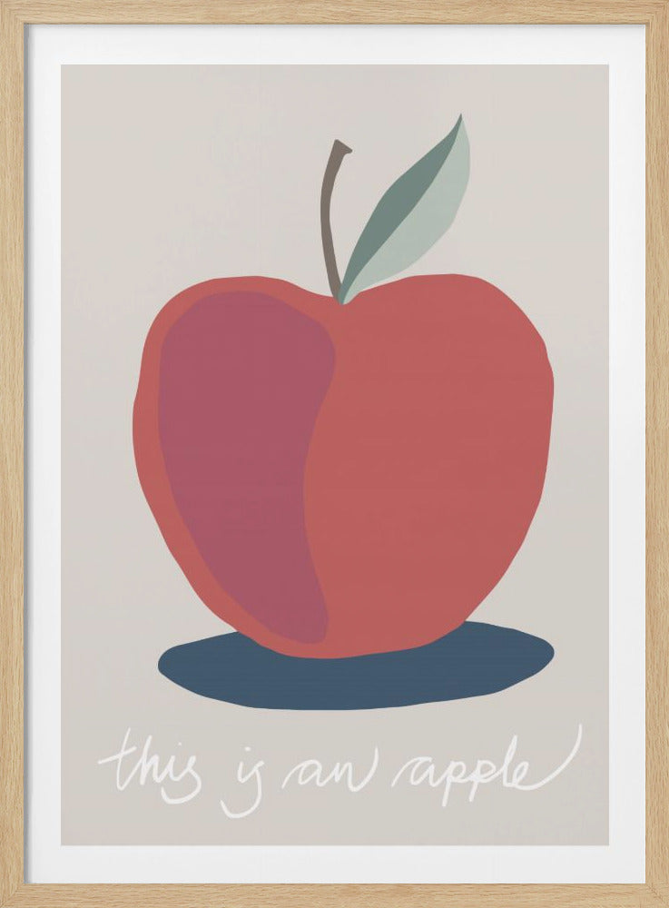 A framed art print featuring a stylized illustration of a red apple on a light beige background. The apple has a single green leaf and casts a blue shadow. Below the fruit, the phrase 'this is an apple' is written in a whimsical white script. Artwork