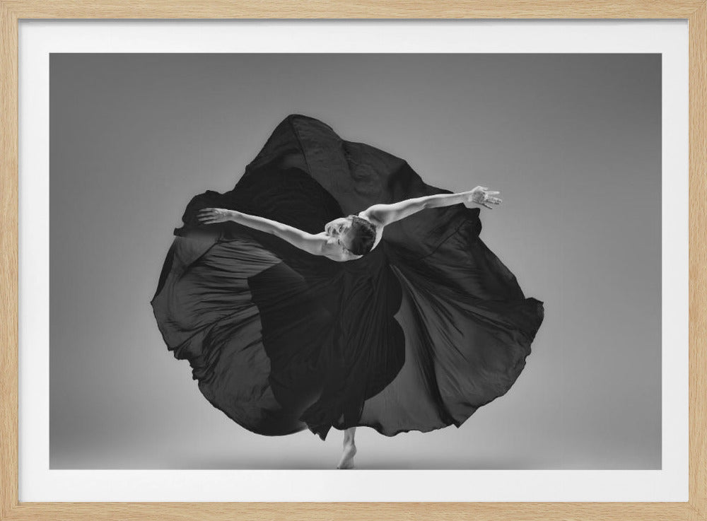 A dramatic black and white photograph of a ballerina in a dynamic pose. Her voluminous black dress billows out around her as she balances gracefully on one toe with her arms outstretched against a simple grey background. The image is shown within a silver frame. Poster