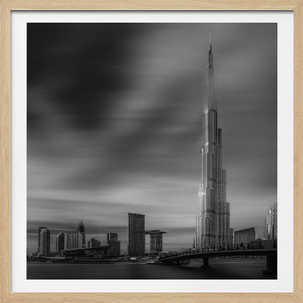 A framed, long-exposure black and white photograph of the Dubai skyline, featuring the iconic Burj Khalifa. The sky has a dramatic, streaky appearance from the long exposure, contrasting with the sharp details of the modern architecture. A bridge crosses the water in the foreground. Print