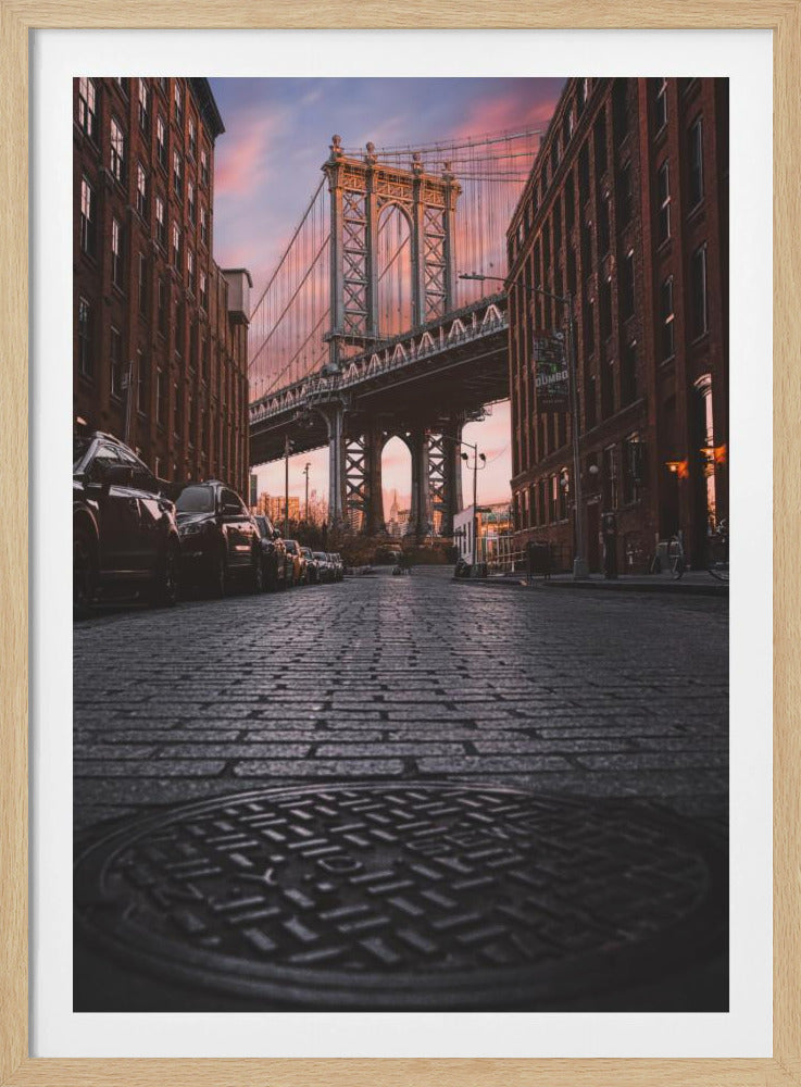 A low-angle photograph taken from a cobblestone street in Dumbo, Brooklyn, looking up towards the Manhattan Bridge at sunset. Red brick buildings frame the view on both sides of the street, with parked cars lining the left. A manhole cover is prominent in the foreground, and the sky is filled with warm pink and orange tones. Print