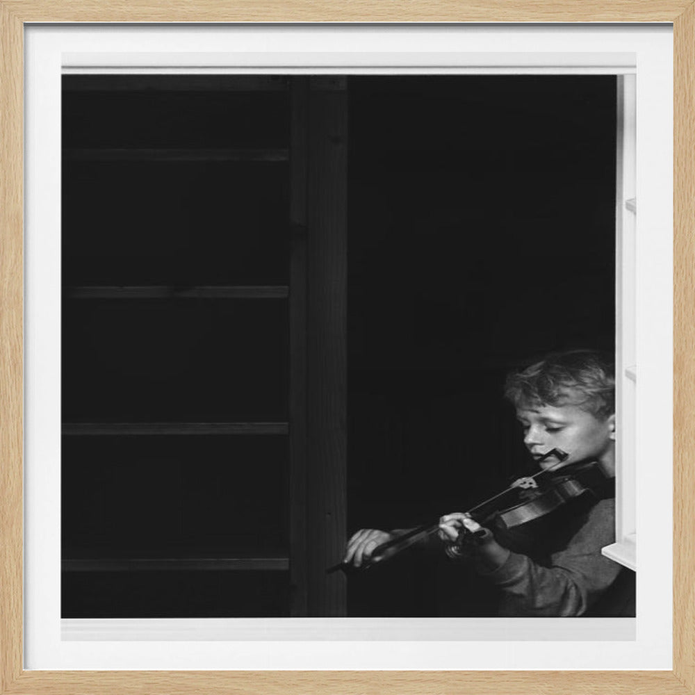 A framed, black and white photograph of a young boy intently playing the violin next to an open window. The dark interior contrasts with the light from the window, highlighting the child's concentration. The image is presented within a light wood frame. Artwork