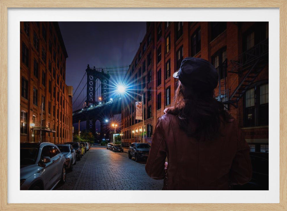 A woman from behind in a brown leather jacket and a black cap stands on a cobblestone street at night in DUMBO, Brooklyn, looking towards the illuminated Manhattan Bridge. The street is lined with brick buildings and parked cars, and a bright streetlight creates a starburst effect. Print