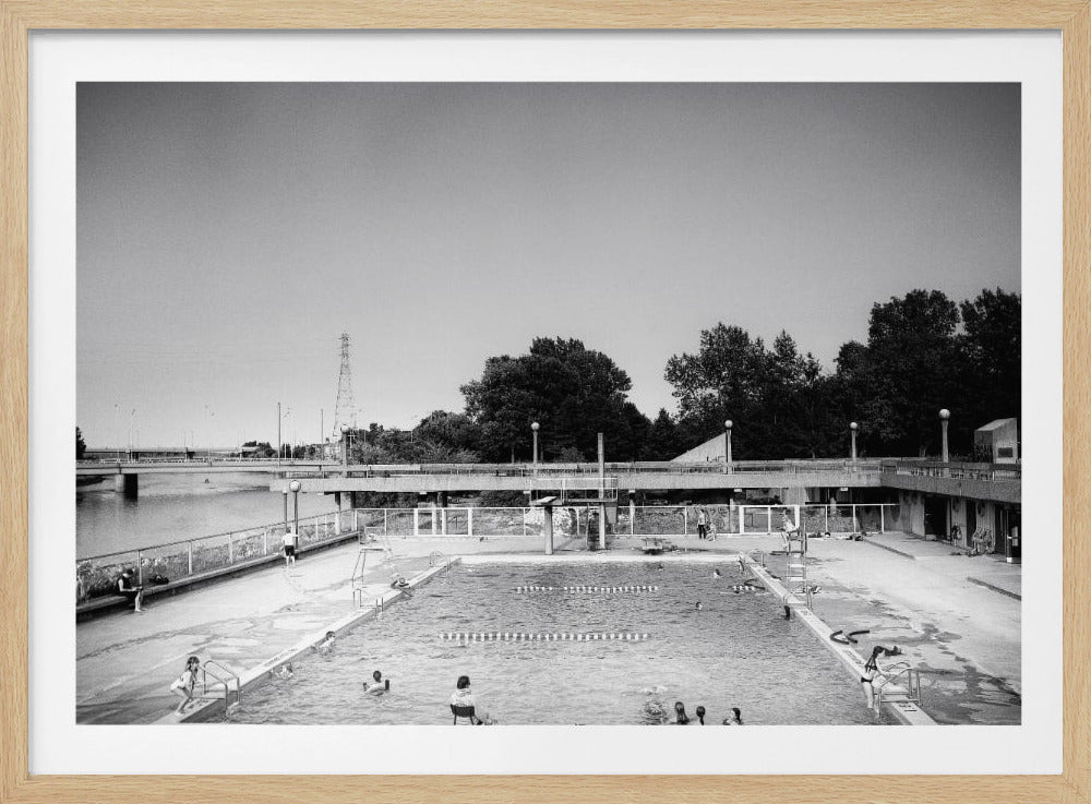 A framed, black and white photograph from a high angle showing a public swimming pool on a summer day. A few people are scattered in the water and sitting on the concrete deck. In the background, a bridge crosses a body of water, with trees and a radio tower visible beyond. Wall Art