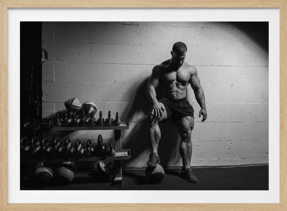 A dramatic black and white photograph of a shirtless, muscular bodybuilder posing in a gym. He leans against a cinder block wall with one foot propped on a medicine ball, his chiseled physique and defined muscles highlighted by intense lighting. Racks of kettlebells are visible in the background. Wall Art