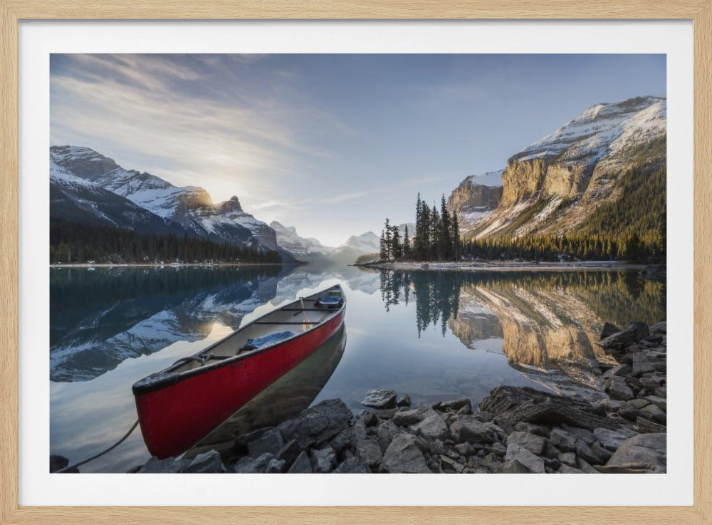 A vibrant red canoe rests on the rocky shore of a calm, glassy lake that perfectly reflects the surrounding snow-capped mountains and a bright, sun-kissed sky. Decor