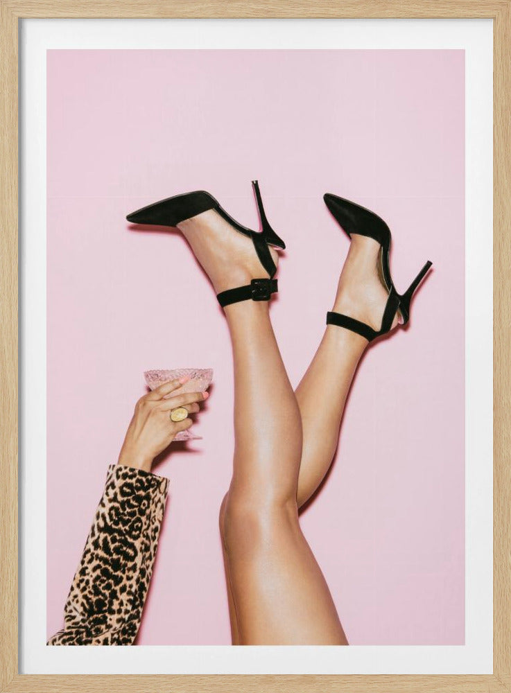 A woman's legs are kicked up in the air against a solid pink background. She is wearing black, pointed-toe, high-heeled shoes with ankle straps. A hand in a leopard print sleeve holds a pink, textured cocktail glass to the side. Artwork