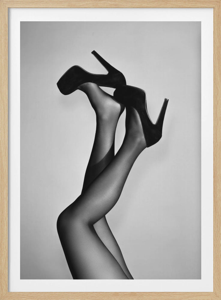 A black and white studio photograph of a woman's legs crossed and raised up, wearing sheer black stockings and black suede platform stiletto high heels against a light gray background, enclosed in a black frame. Wall Art