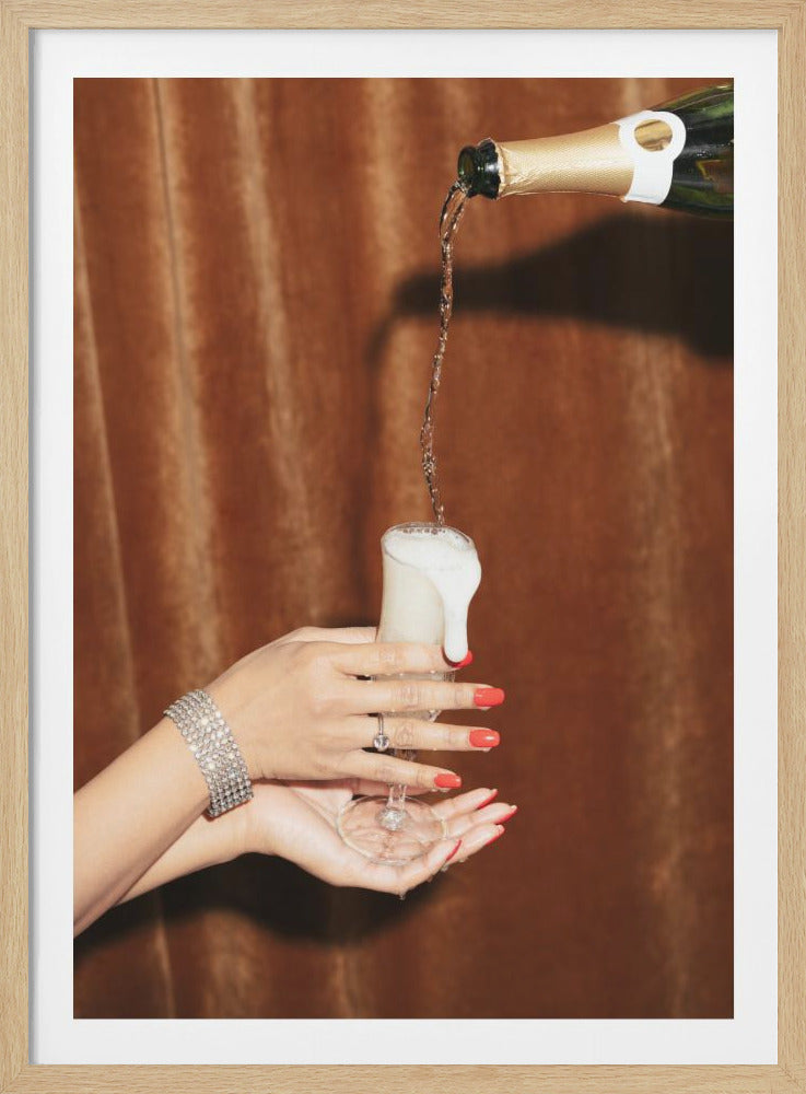 A close-up shot of champagne being poured from a gold-foiled bottle into a flute glass, causing it to overflow with white foam. The glass is held by a woman's hands with vibrant red nail polish, a sparkling diamond bracelet, and a ring, set against a rich brown velvet curtain. Decor