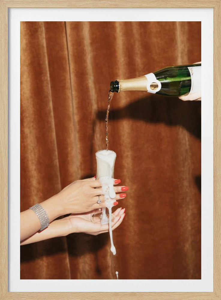 Champagne is poured from a bottle into a glass, overflowing with white foam that drips down onto two manicured hands with red nail polish holding it against a brown velvet curtain background. Poster