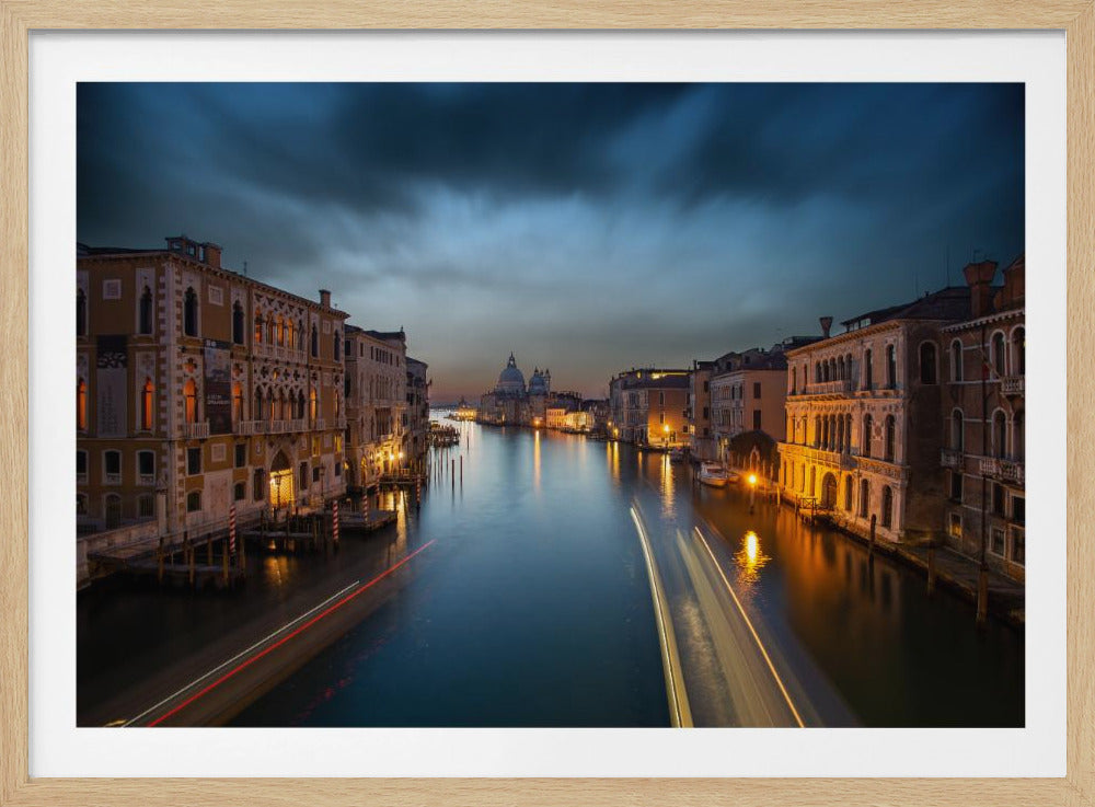 A long-exposure photograph of the Grand Canal in Venice at dusk, framed in silver. Ornate, illuminated buildings line the canal, leading to the distant dome of Santa Maria della Salute under dramatic dark blue clouds. Streaks of light from moving boats are visible on the calm water. Wall Art