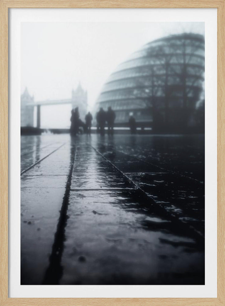 A moody, black and white, low-angle photograph of a wet city pavement in London. The foreground shows shimmering reflections on the dark, wet ground. In the blurry, foggy background, silhouettes of people stand near the modern City Hall building, with the iconic Tower Bridge visible further in the distance. Decor