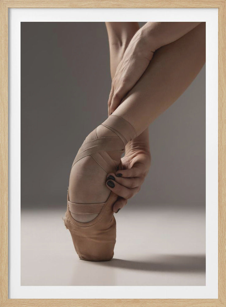 A close-up artistic photograph of a ballerina's foot in a beige pointe shoe, standing on the tip of the toe. Two hands with black nail polish gently hold the ankle, set against a neutral gray background within a black frame. Decor