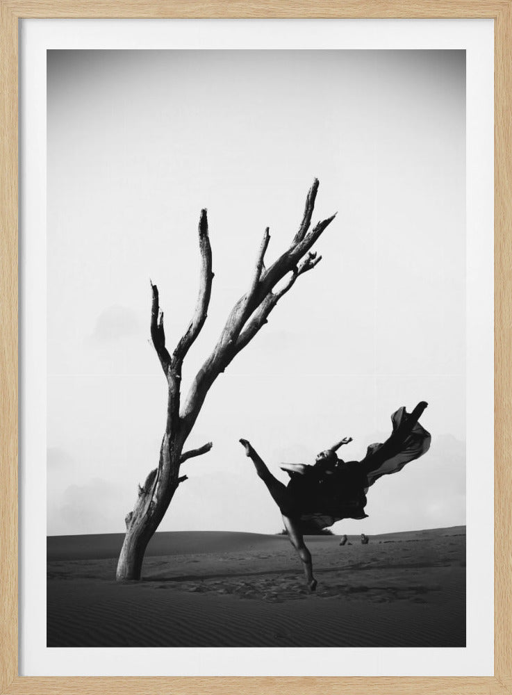 A dramatic black and white photograph of a female dancer in a dark, flowing dress, captured mid-leap next to a gnarled, dead tree in a barren desert landscape. Poster
