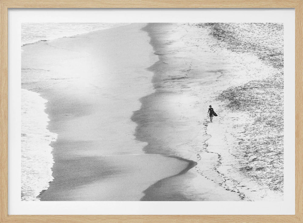 A high-angle, black and white photograph of a lone surfer walking along a sandy beach with a surfboard under their arm. The image has a minimalist feel, focusing on the patterns of the wet and dry sand and the single figure against the vastness of the shore. The artwork is presented in a silver frame. Artwork
