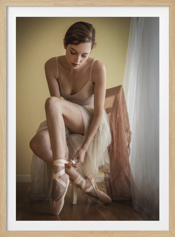 A ballerina with brown hair pulled back sits on a white chair, looking down as she ties the pink ribbons of her pointe shoes. She is wearing a cream-colored leotard and a sheer, light-colored skirt over it. The background is a soft yellow wall with a sheer white curtain to the right, and the floor is made of wood. Wall Art