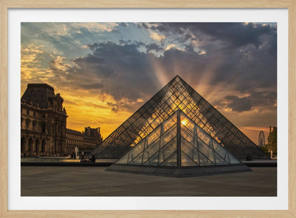 A striking photograph of the glass Louvre Pyramid at sunset, with the sun's rays bursting from behind its peak into a dramatic, cloudy sky. The golden light of the sunset is reflected in the pyramid's glass panels, contrasting with the dark silhouettes of the surrounding historic buildings. Print