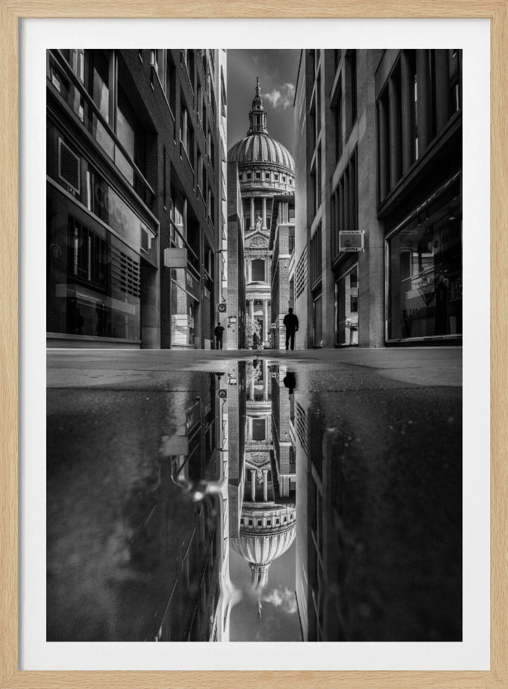 A dramatic black and white photograph taken from a low angle on a narrow city street, perfectly framing St. Paul's Cathedral in the distance. The foreground is dominated by a large puddle, which creates a stunning, clear reflection of the entire scene, including the cathedral, buildings, and sky. Print