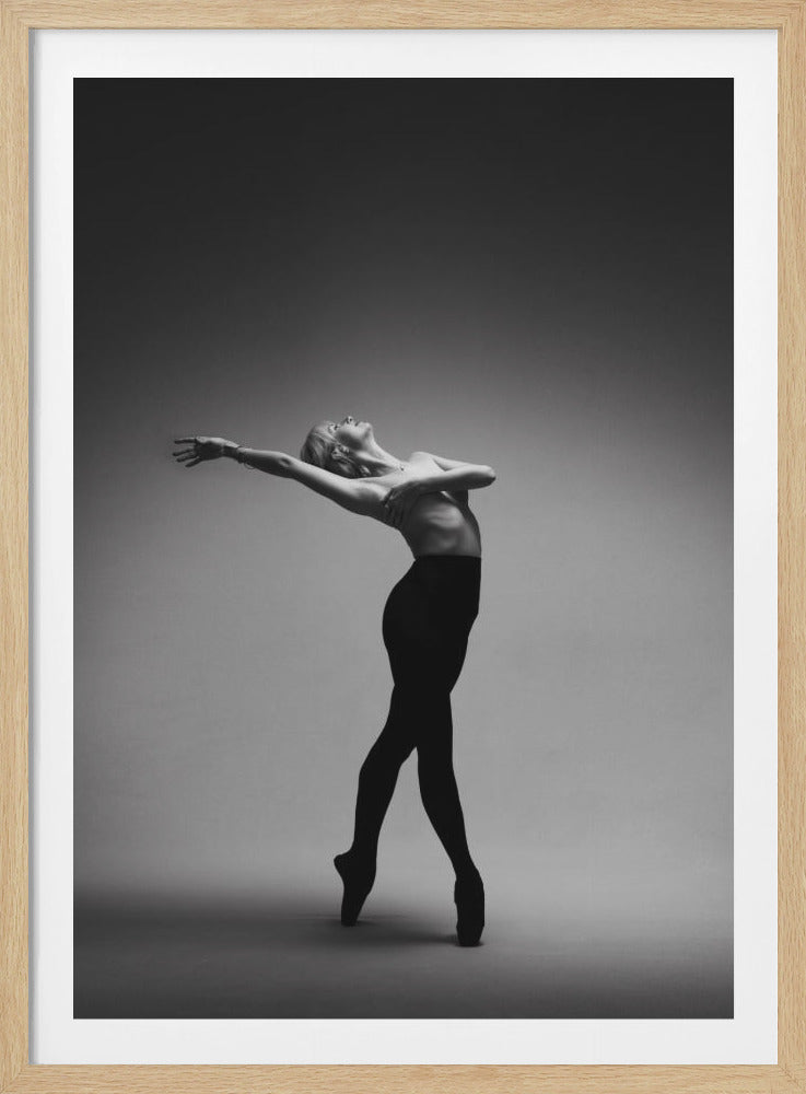 A dramatic black and white photograph of a ballerina in a graceful pose against a simple gray background. She stands en pointe in black tights, her upper body bare, with her arms elegantly positioned and her head tilted back in an expressive arch. Wall Art
