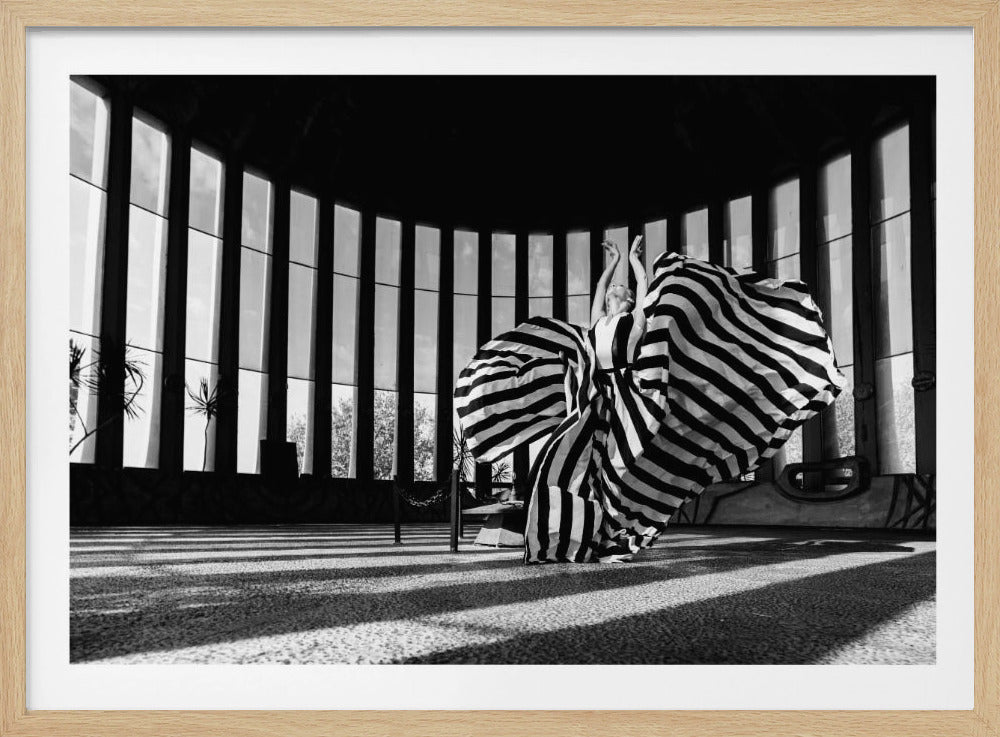 A dramatic black and white photograph of a woman in a voluminous, horizontally striped dress, captured mid-motion with the fabric swirling around her. She stands in a sunlit, circular room with tall, slatted windows that cast long shadows across the floor. Her arms are raised gracefully above her head, creating a striking and artistic pose. Artwork