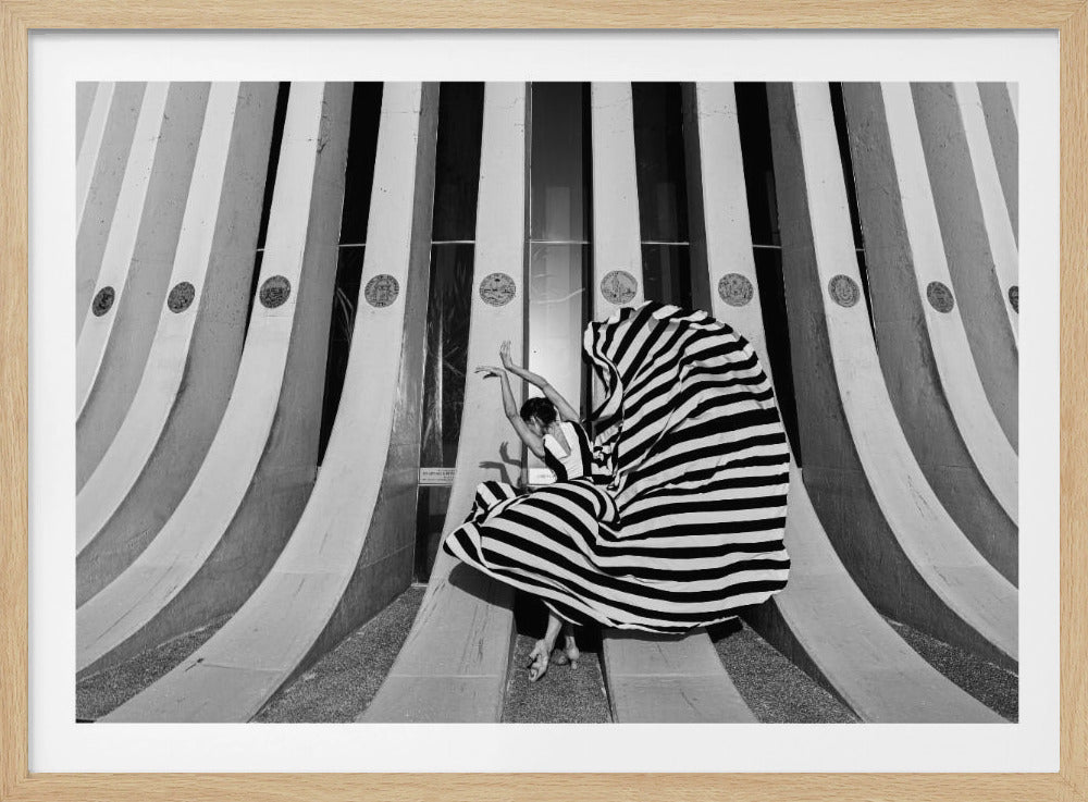 A framed, black and white photograph of a woman in a large, flowing black and white striped dress. She is in a dance pose in front of an architectural structure with large, curved, striped columns that mirror the pattern of her dress, creating a strong graphic and linear composition. Artwork