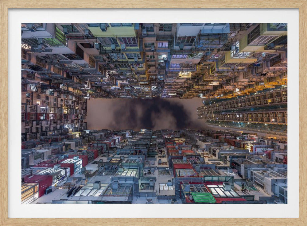 A surreal, symmetrical photograph looking up from the courtyard of a densely packed apartment complex at night. The buildings create an enclosure around a dark, cloudy sky, and the top half of the image is an inverted reflection of the bottom half, creating a disorienting, futuristic cityscape. The scene is illuminated by the glow of numerous windows and is presented within a silver picture frame. Artwork