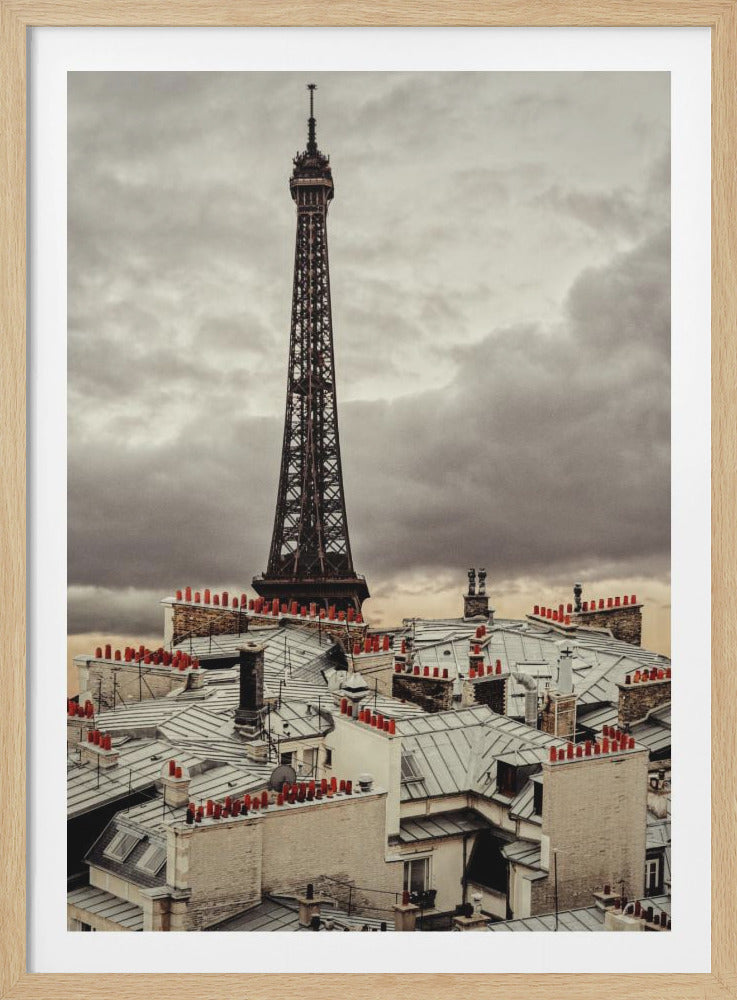A dramatic, high-angle view of the Eiffel Tower in Paris, France, looming over a dense cluster of traditional Parisian rooftops under a heavy, overcast sky. The muted tones of the scene are punctuated by the bright red of numerous chimney pots. Print