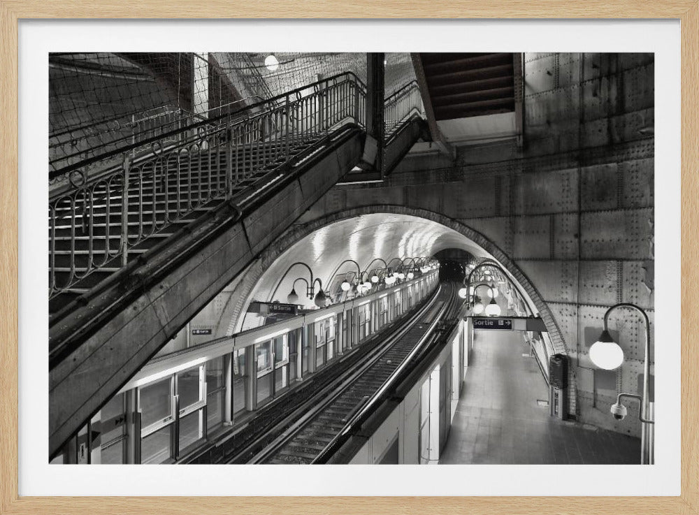 A high-angle, black and white photograph of an empty, vintage-style subway station. Ornate stairs are on the left, leading up from the platform below. The tracks curve into a dark, arched tunnel, and the platform is illuminated by a series of round, curved lamps. A sign with the word "Sortie" is visible on the platform. Artwork