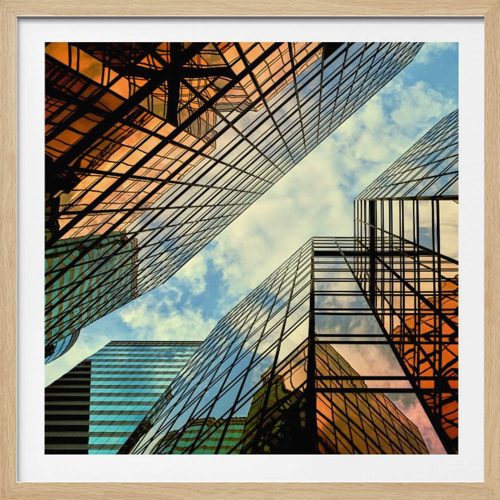 A low-angle photograph looking up at the converging facades of several modern glass skyscrapers, which reflect a blue sky with white clouds and a brilliant orange sunset. The image is enclosed in a light wood frame. Decor