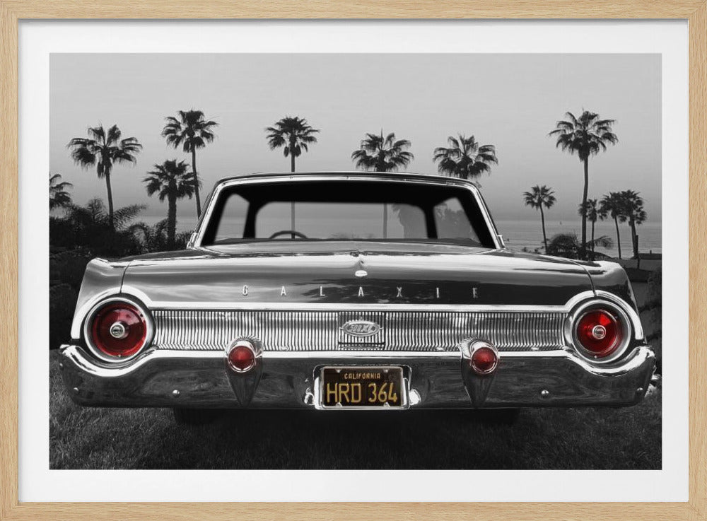 A selective color, black and white photograph of the rear of a vintage Ford Galaxie. The car is parked on grass with a row of palm trees and the ocean in the background. Only the round red taillights and the yellow California license plate reading "HRD 364" are in color, contrasting with the monochrome scene. The entire image is presented within a silver frame. Print