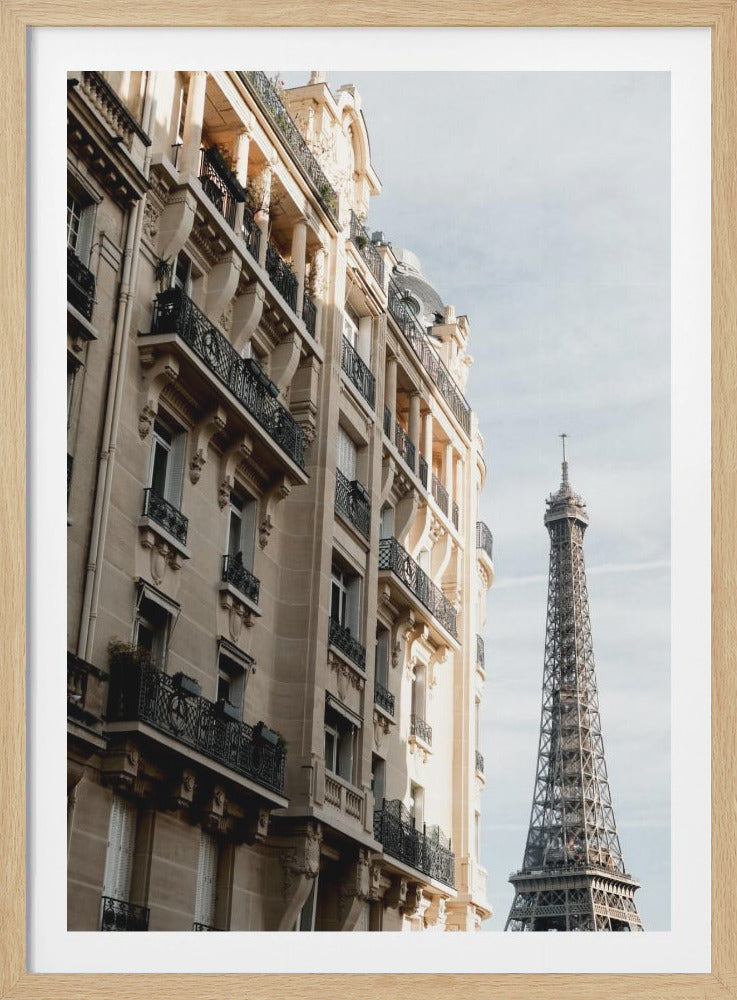 A low-angle photograph of a classic beige Parisian apartment building with intricate black wrought-iron balconies, with the iconic Eiffel Tower visible in the background against a pale, hazy sky. Print