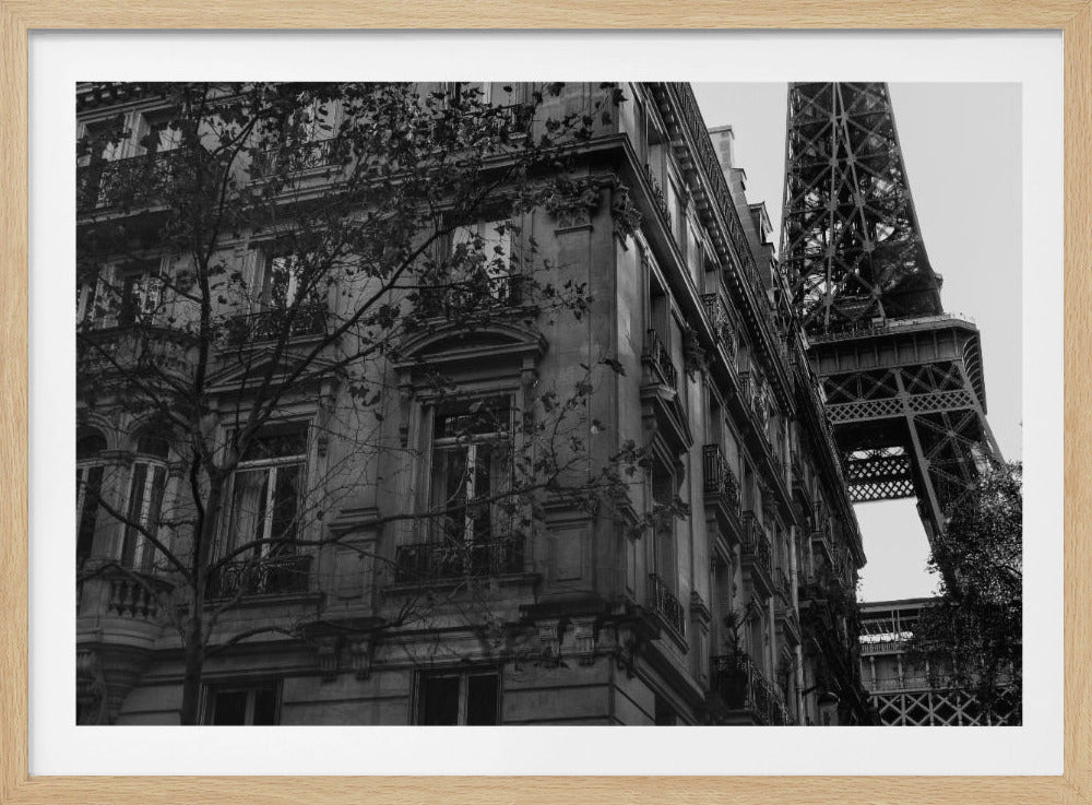 A framed, black and white photograph showing a low-angle view of an ornate Parisian apartment building, with the top of the Eiffel Tower peeking out from behind it against a light sky. Poster
