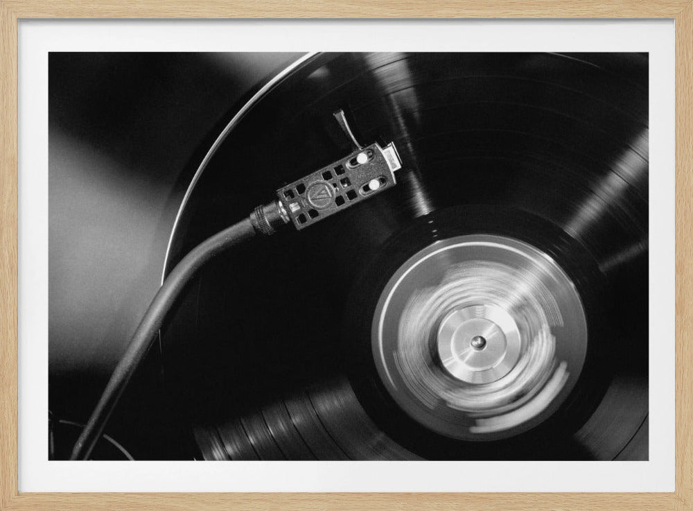 A framed, black and white, close-up photograph of a record player in action. The turntable is spinning a vinyl record, creating a motion blur on the center label. The tonearm and stylus are positioned over the grooved surface of the record. Poster