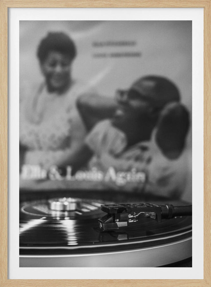 A black and white photograph focusing on a turntable's stylus resting on a spinning vinyl record. In the blurred background, the album cover for 'Ella & Louis Again' featuring Ella Fitzgerald and Louis Armstrong is visible. Poster