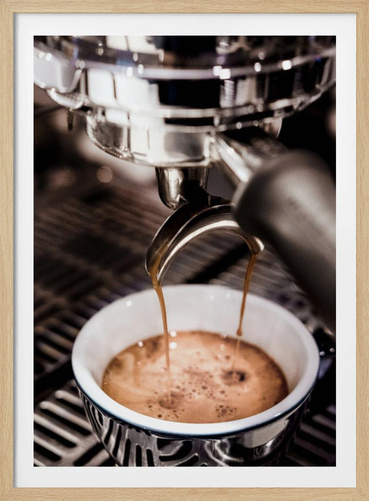 A close-up photograph of a shiny chrome espresso machine brewing a fresh shot of espresso. Two streams of rich, dark coffee pour from the portafilter into a white ceramic cup with a blue patterned rim, creating a light brown crema on top. Print