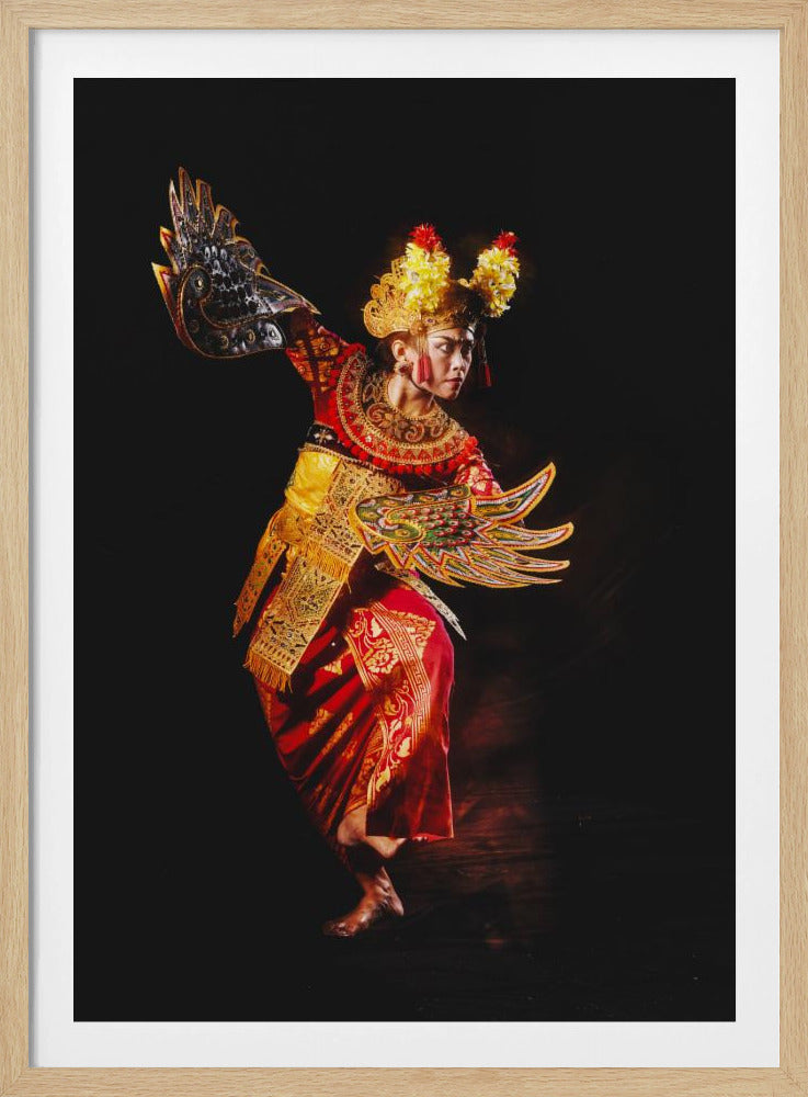 A full-length studio portrait of a female Balinese dancer mid-pose against a solid black background. She is dressed in a traditional, ornate costume of red and gold, including an elaborate headdress with yellow and red flowers and wing-like accessories on her arms. The lighting is dramatic, highlighting the vibrant colors and the dancer's intense, focused expression. Poster