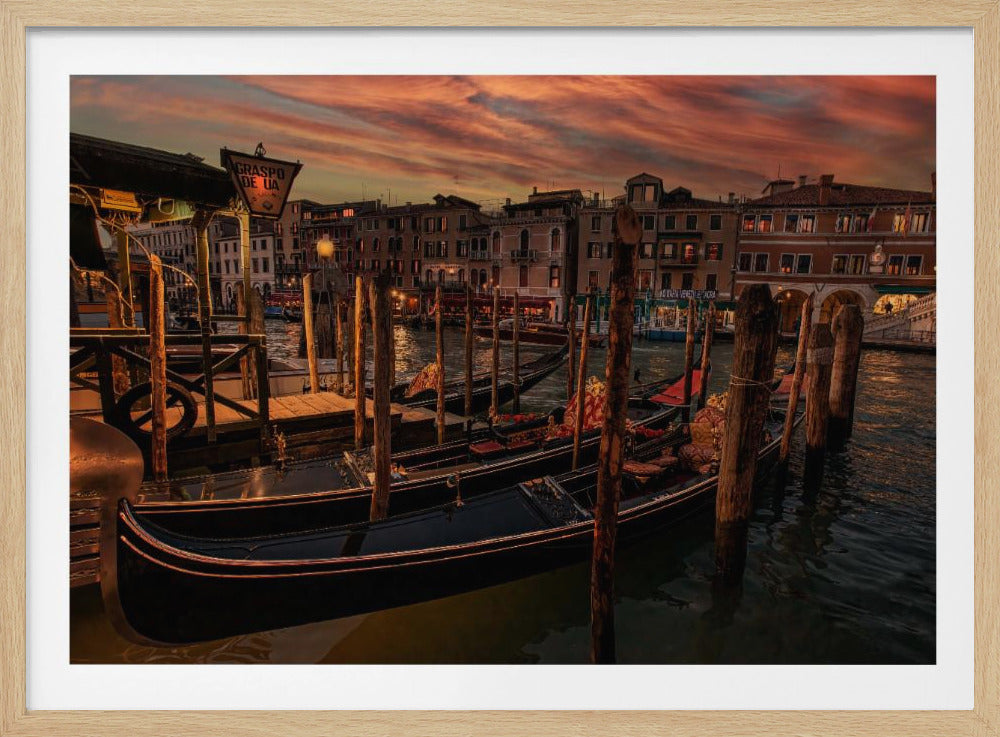 A romantic view of gondolas docked in a Venice canal at sunset. The sky is a dramatic mix of orange and red, reflecting on the water, while the lights from the historic buildings along the canal begin to glow. Print