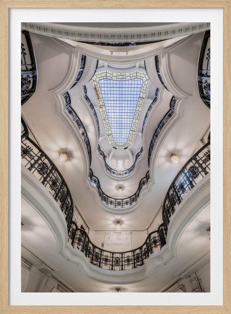 A low-angle, worm's-eye view looking straight up through the multiple floors of a grand, oval-shaped atrium. Ornate, black wrought-iron railings create sweeping curves against the bright white walls and ceilings, leading the eye towards a large, grid-patterned glass skylight revealing a blue sky. Print