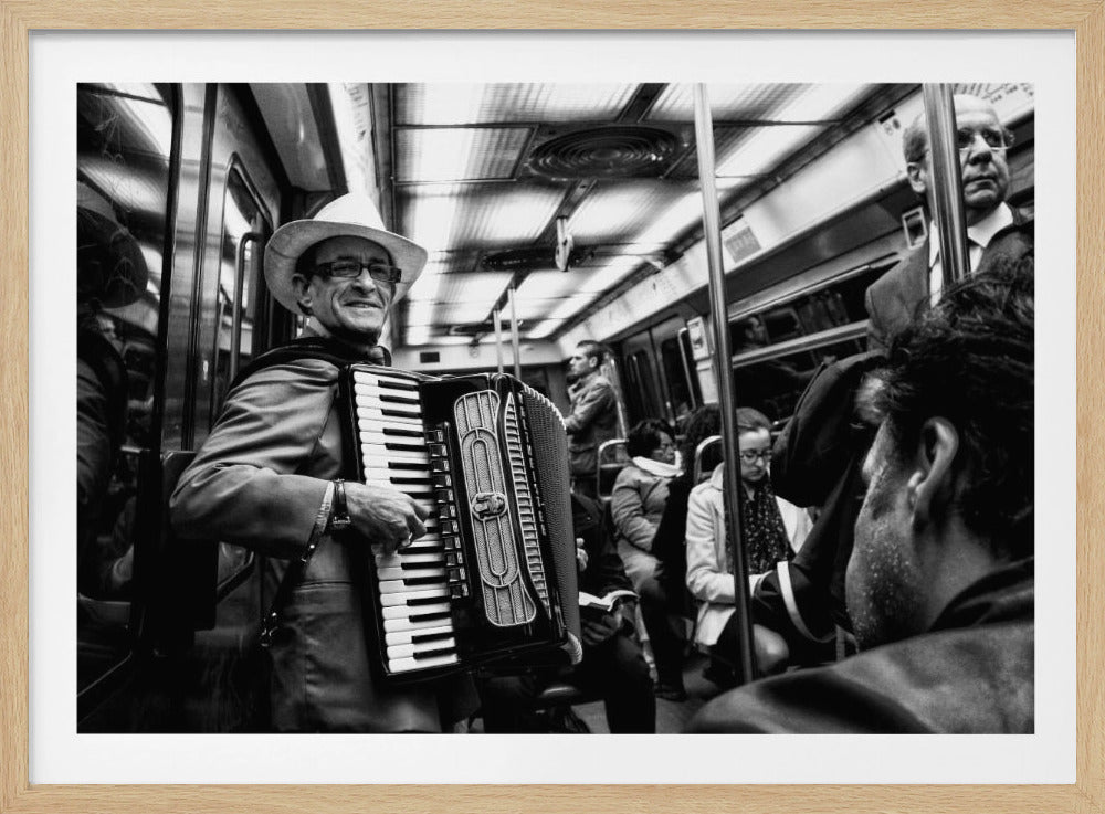 A black and white photograph of a smiling man in a fedora playing an accordion for fellow passengers on a crowded subway train. Print