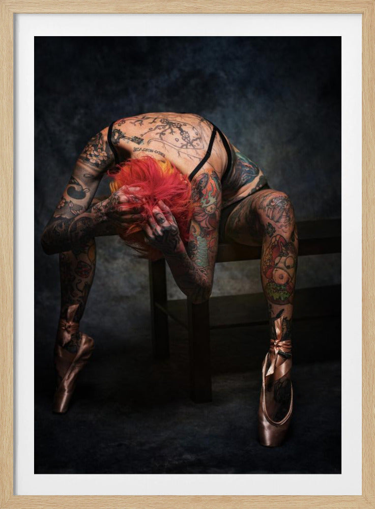 A heavily tattooed ballerina with vibrant red hair performs an extreme backbend while seated on a dark bench. She is wearing black lingerie and pink satin pointe shoes, with her face obscured as her hands clutch her head. The background is a dark, moody gray. Poster