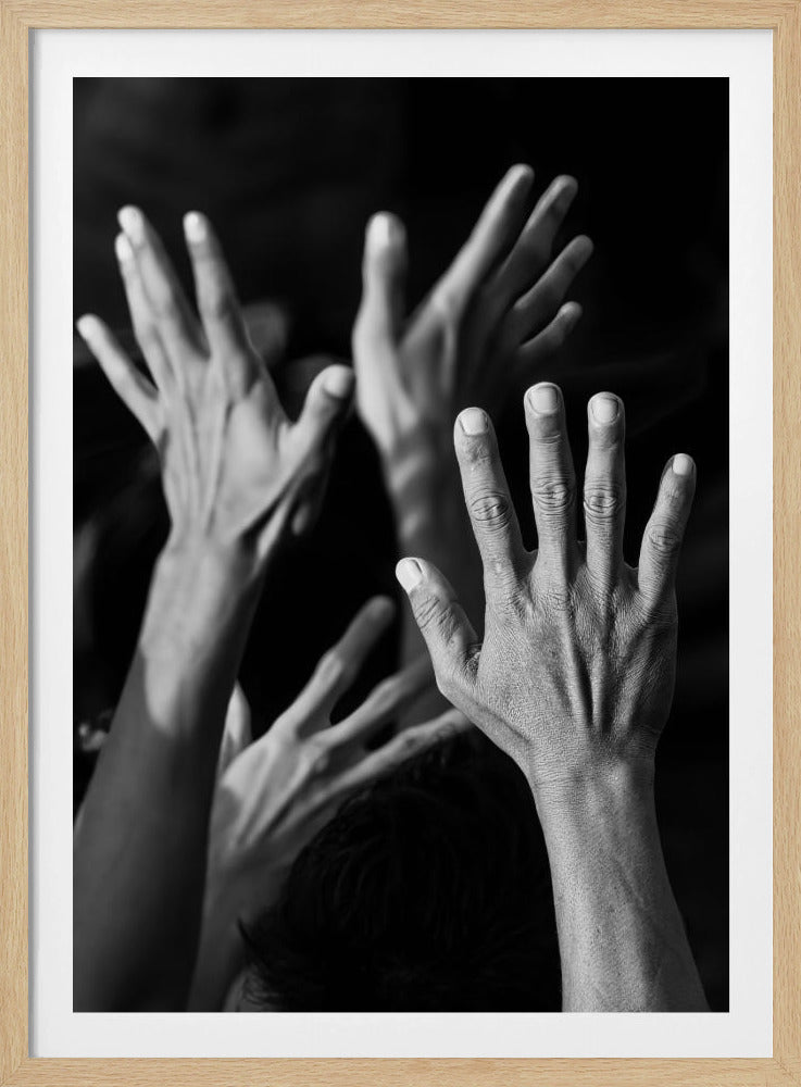 A dramatic black and white photograph capturing multiple hands reaching upwards against a dark background. A single hand in the foreground is sharply detailed, its palm open and fingers spread, while other hands fade into the soft-focus background, creating a powerful sense of unity and expression. Artwork